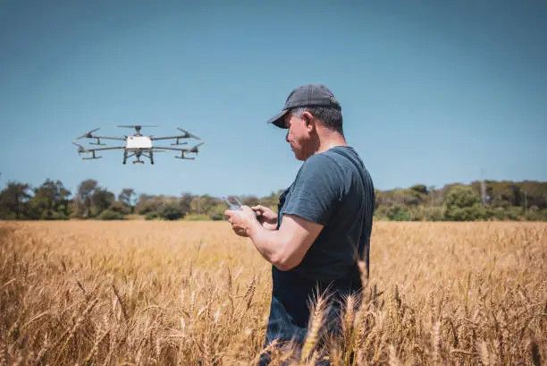 A farmer monitoring a drone sprayer via a tablet, with a field of crops in the background - Agristuff